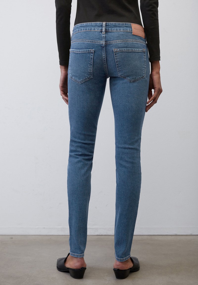 Chude niebieskie dżinsy z wysokim stanem, pięcioma kieszeniami i brązową skórzaną metką z logo na tylnej części pasa, prezentowane na figurze.