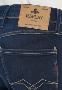 Σκούρο μπλε τζιν παντελόνι διαθέτει ταμπέλα από καφέ δέρμα με ανάγλυφο το όνομα "REPLAY", χρυσή ραφή και διακοσμητικό λευκό ζιγκ ζαγκ σχέδιο στις τσέπες.