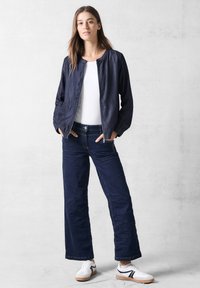 Dunkelblaue Bomberjacke mit Rundhalsausschnitt, kombiniert mit passenden locker sitzenden dunklen Hosen und weißen Sneakers mit schwarzen Akzenten.