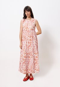 Maxikleid aus weißem Stoff mit kunstvollen orangen Mustern, ärmelloses Design und geraffter Taille. Kombiniert mit roten Ballerinas.