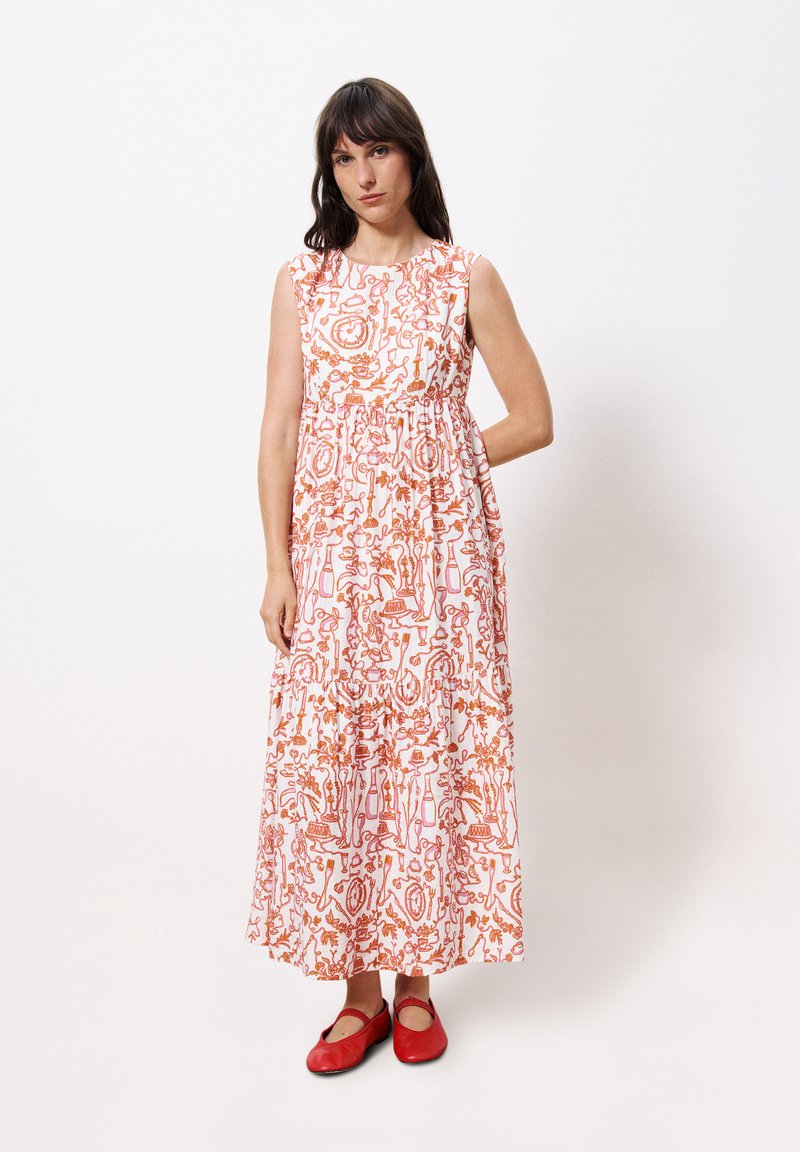 Maxikleid aus weißem Stoff mit kunstvollen orangen Mustern, ärmelloses Design und geraffter Taille. Kombiniert mit roten Ballerinas.