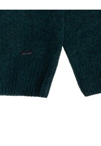Gebreid sweater in donker teal, met een geribbelde textuur en een V-vormige zoom. Klein logo-accent in zwart en rood nabij de zoom.