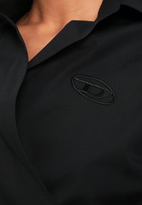 Μαύρο πουκάμισο με γιακά, λεία υφή και κεντημένο λογότυπο στην αριστερή πλευρά του στήθους. Το ύφασμα φαίνεται ελαφρύ.