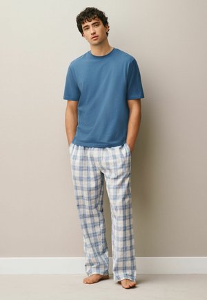 Junger Mann steht barfuß, trägt ein blaues Kurzarmhemd und weiße und blaue karierte Pyjamahosen vor einer einfarbigen beigen Wand.