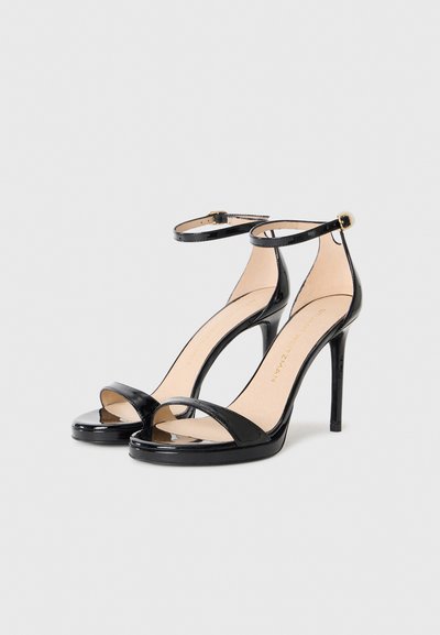 Sandales à talons hauts en cuir verni noir avec des brides autour de la cheville et des orteils ouverts, placées côte à côte sur un fond blanc.