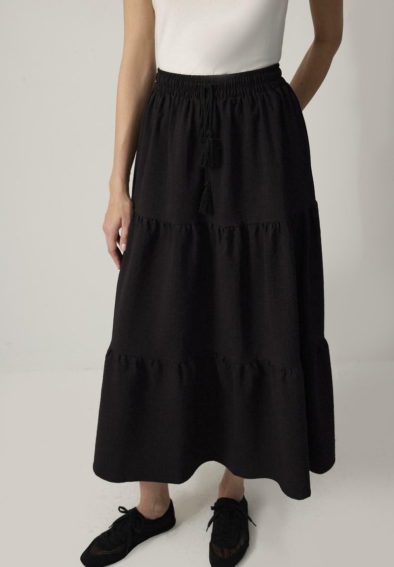 Γυναίκα που φοράει μακριά μαύρη φουστάνια με στρώσεις και ελαστική μέση με κορδόνι, σε συνδυασμό με μαύρα παπούτσια με κορδόνια και λευκό τοπ.