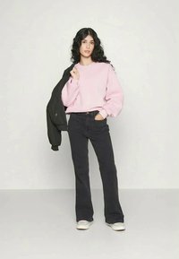 Ροζ oversized φούτερ, μαύρα flare τζιν, μαύρο μπουφάν ριγμένο στον ώμο, λευκά αθλητικά παπούτσια, μακριά καστανά μαλλιά, ουδέτερο φόντο.