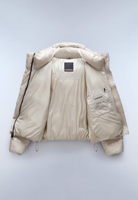 Giacca a vento beige con finitura lucida, dotata di un ampio cappuccio, chiusura con zip e tasche. L'interno include un'etichetta e una tasca di stoccaggio.