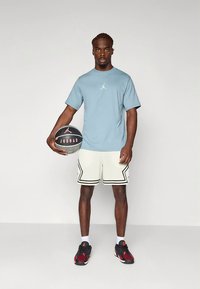 Blå bomull t-skjorte med logo, hvite perforerte shorts med svarte detaljer, og røde/svarte joggesko. Modellen holder en basketball.