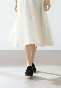 Sabots en daim noir avec des clous en métal sur la semelle, associés à une robe blanche fluide et transparente avec une finition texturée et un ourlet effiloché.