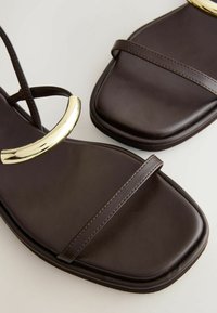 Sandales en cuir marron foncé avec des bouts carrés, des fines brides, et un détail en métal doré incurvé sur la bride de la cheville.