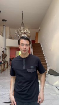 Jeune homme aux cheveux foncés portant une chemise noire, debout dans une pièce moderne et spacieuse avec un lustre et un escalier en bois en arrière-plan.