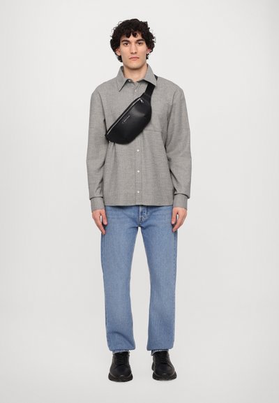 Un sac bandoulière noir porté sur une chemise à boutons grise et un jean bleu. La chemise possède une texture douce, et le jean a une coupe droite.