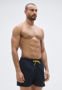 Tengerészkék fürdőnadrág sárga zsinórral, sima textúrával és rugalmas derékrésszel. Logó részlet az elején.