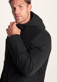 Man adjusting the collar of a black hooded jacket with a small logo on the upper sleeve against a plain light background.