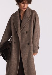 Brązowy wełniany płaszcz dwurzędowy z dużymi klapami, zapięciem na dwa guziki i bocznymi kieszeniami; model trzymający dopasowany materiał.