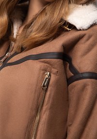Veste en faux suede marron avec un col en fausse fourrure crème, des accents noirs et un zip doré sur la poche latérale.