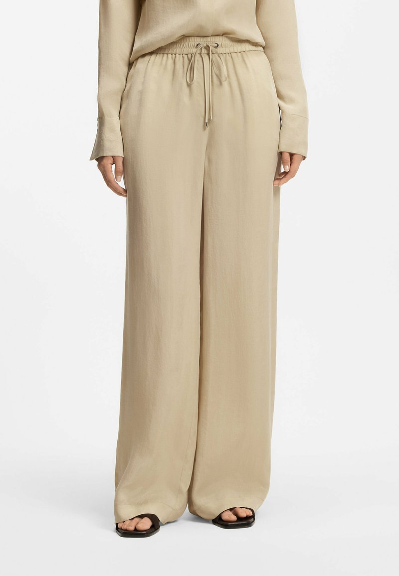 Pantalon beige à jambes larges avec taille élastique et cordon de serrage, porté avec un haut à manches longues beige assorti et des sandales noires à bout carré.