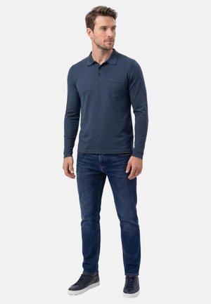 Man staat en draagt een marineblauw poloshirt met lange mouwen, donkerblauwe spijkerbroek en zwarte sneakers met witte zolen tegen een effen achtergrond.