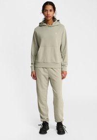 Haut à capuche vert olive clair avec une poche kangourou, des poignets et un ourlet côtelés. Associé à un pantalon de jogging assorti, complété par des chaussures de sport noires.