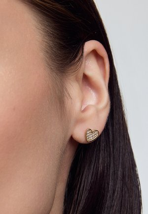 Złote serduszka w kształcie kolczyków z przezroczystymi cyrkoniami, umieszczone na zbliżeniu ucha i ciemnych włosów. Gładka faktura, mały rozmiar.