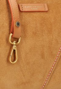 Beige suède handtas detail met een tan leren schouderband, gouden hardware sluiting en een gestikt accent in het rood. Merkenlabel zichtbaar.