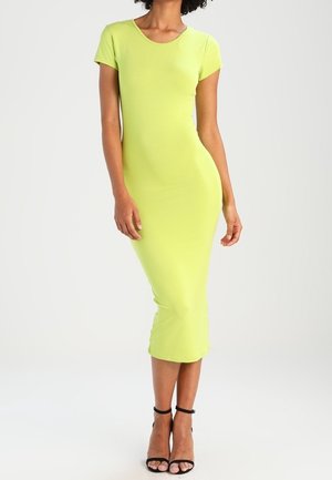 Femme portant une robe midi ajustée à manches courtes vert citron et des sandales noires à talons hauts avec bride à la cheville, debout devant un fond uni.