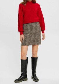 Žena v červeném svetru, kostkované sukni a černých vysokých kozačkách stojící na jednolitém pozadí.