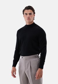 Schwarzer Strick-Pullover mit Rollkragen und langen Ärmeln, kombiniert mit hellgrauen, plissierten Hosen, die über einen Knopf- und Riemenverschluss verfügen.