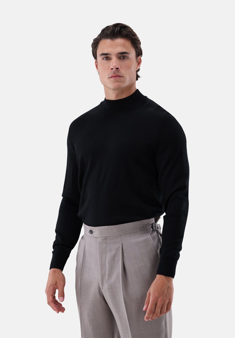 Schwarzer Strick-Pullover mit Rollkragen und langen Ärmeln, kombiniert mit hellgrauen, plissierten Hosen, die über einen Knopf- und Riemenverschluss verfügen.