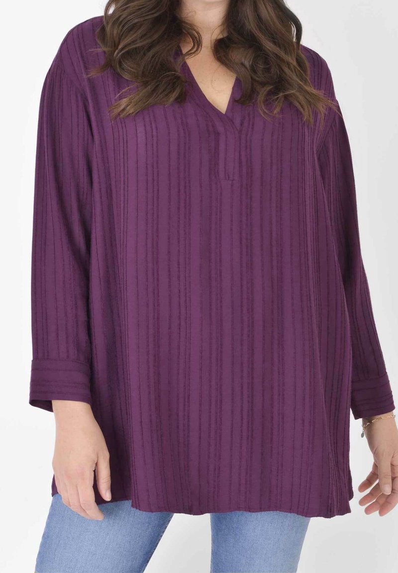 Maglietta viola a maniche lunghe con scollo a V e texture a coste verticali, caratterizzata da una vestibilità rilassata e polsini arrotolati. Abbinata a jeans azzurri chiari.