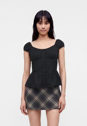 Junge Frau trägt ein schwarzes, kurzärmeliges, geknöpftes Peplum-Top und einen kurzen karierten Rock, steht vor einem schlichten hellen Hintergrund.
