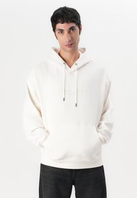 Cremefarbene Kapuzenjacke aus weichem Material, mit einer Vordertasche, verstellbaren Kordeln und dezentem Logo-Stick über der Tasche.