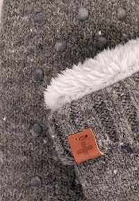 Grijze gebreide beanie met een textuurpatroon van verhoogde stippen, een witte nepbont manchet en een bruin leren label met een kruisdetail.