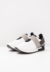 Vita sneakers med en slät yta, som har ett grått elastiskt band, svart hälpanel och en strukturerad svart gummisula med vita accenter.