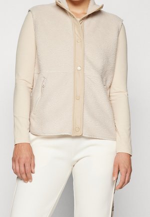 Person iført en beige fleecemodel vest med trykknapper over en langærmet ribbet trøje og hvide bukser med sidedetalje, hænder afslappet ved siderne.