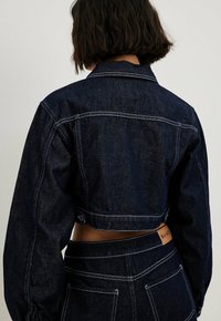 Lyhyt tumma farkkutakki pitkällä hihaisella, jossa on kontrastivärinen ompelu ja takana paikka-label. Takki on muotoiltu istuvasti.