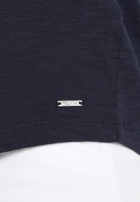 Granatowa koszulka wykonana z lekkiego materiału, z małą srebrną metką. Rękaw jest starannie wykończony i ma teksturowane wykończenie.