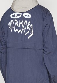 Navy jacket with a light gray hood, featuring white graphic design of eyes and hands on the back. Made of textured, lightweight fabric.
