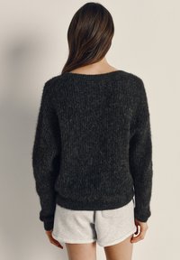 Person mit langen braunen Haaren, die einen dunklen, flauschigen Pullover und hellgraue Shorts trägt, steht mit dem Gesicht zu einer schlichten, elfenbeinfarbenen Wand.