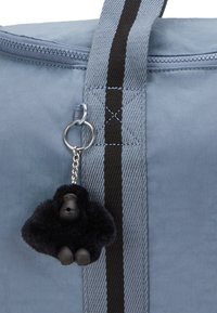 Lys blå taske med en sort stribet rem, der har en nøglering af en sort gorilla plysbamse med et plastikansigt, og lynlås lukning.