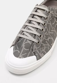 Grijze textielsneaker met een subtiel patroon, witte rubberen neuskap en veters. Voorzien van tonale stiksels en ronde veterogen.