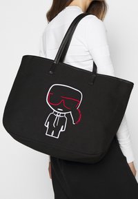 Svart canvas-tygväska med svarta läderremmar. Har ett broderat motiv i vitt och rött som tillför ett lekfullt designelement.