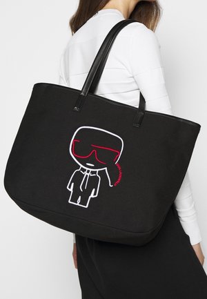 Zwarte canvas tote bag met zwarte leren handvatten, met een wit en rood geborduurd cartoonfiguur op de voorkant. Grote, rechthoekige vorm.