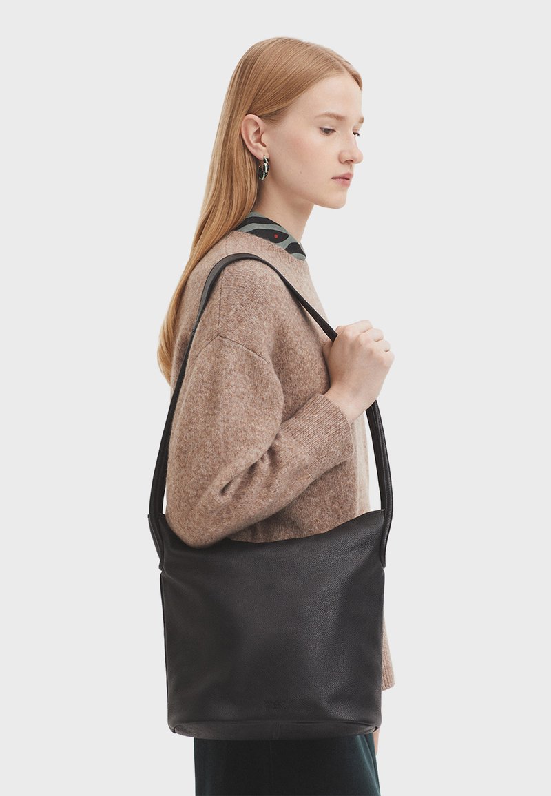 Sac à épaule en cuir noir avec une texture lisse et une large bandoulière, présentant un design simple et spacieux. Le modèle tient le sac devant un fond clair.