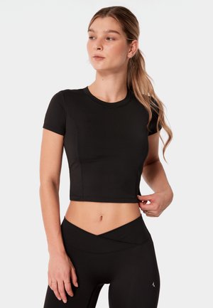 Kobieta o długich blond włosach, ubrana w czarny krótki top z krótkim rękawem i legginsy z wysokim stanem, pozująca na jednolitym jasnym tle.