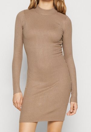 Femme portant une robe en maille brun clair, moulante, à manches longues côtelées et col montant, debout devant un fond clair uni.