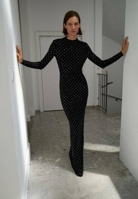 Robe noire ajustée avec manches longues, ornée de pois blancs, atteignant le sol. Texture lisse, design minimaliste.