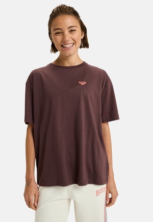 Une jeune femme souriante portant un t-shirt noir violet ample avec un petit logo et un pantalon clair avec le texte "ROXY" sur la cuisse gauche.