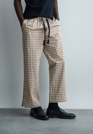 Mężczyzna ubrany w beżowo-białe, szerokie spodnie w kratę z czarnymi sznurkami, czarne mokasyny i ciemny, bezrękawnik, stojący w pomieszczeniu.